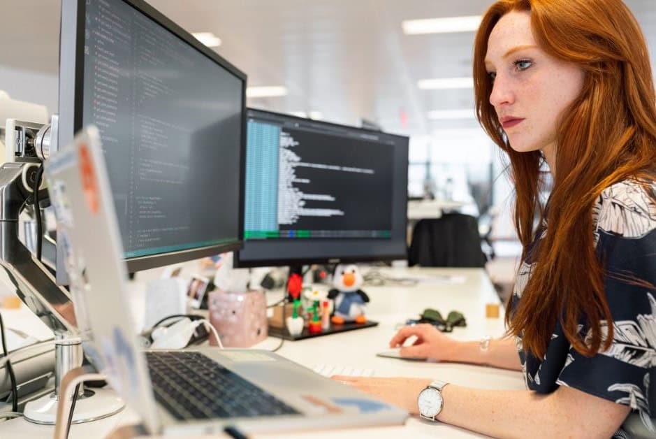 A red haired white woman who is a Blockchain Developer sitting at a desk with two computer screens and a laptop.