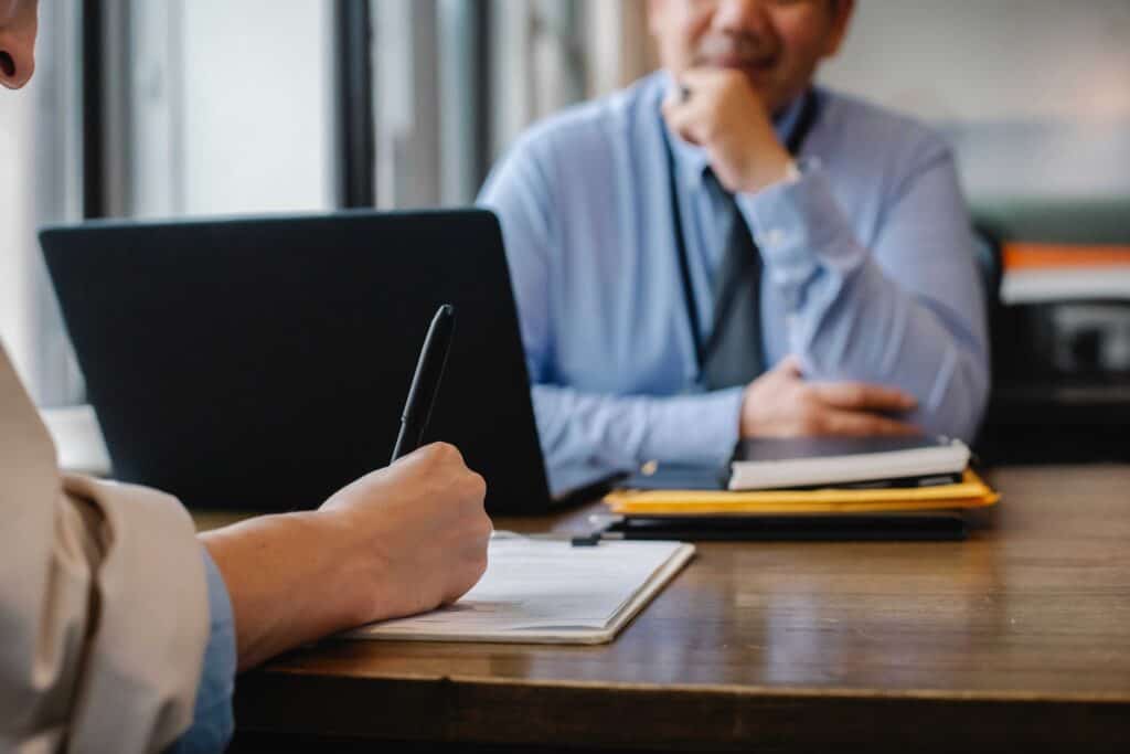 An image of a person writing notes on a clipboard and another man behind a computer. One is drafting his resume manually and the other has already completed his resume with an AI Resume Builder.  He is waiting and pondering why the other person didn't use an AI Resume Builder.