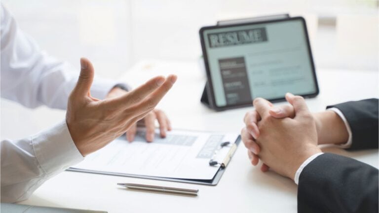 A man is talking to another man at an interview. In the background is a tablet with an image of a resume on the screen built by an AI Resume Builder.