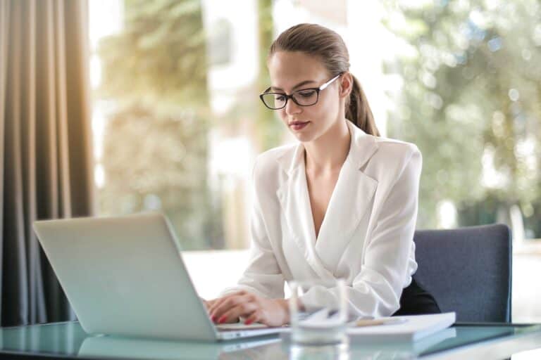 Picture of an AI Engineer working on her computer. It is a lady with a white suit, wearing glasses and her hair is in a pony tale.